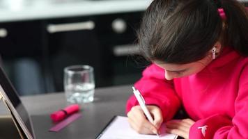 junge Frau in einem hellrosa Sweatshirt sitzt an einem Tisch mit Tablet-Ohrhörern, während sie mit einem Stift in ein Notizbuch schreibt
