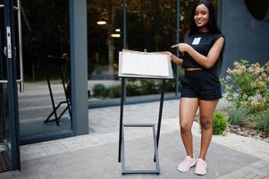 Fashionable feminist african american woman wear in black t-shirt and shorts, posed outdoor and show menu of restaurant. photo