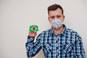 el hombre con camisa a cuadros muestra la tarjeta de la bandera de brasil en la mano, usa una máscara protectora aislada en el fondo blanco. concepto de coronavirus de los países americanos. foto