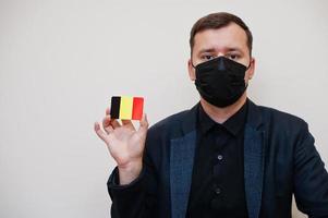 European man wear black formal and protect face mask, hold Belgium flag card isolated on white background. Europe coronavirus Covid country concept. photo