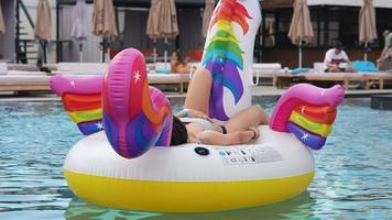 Young woman in a bikini rests on a big rainbow unicorn float while others lounge poolside video