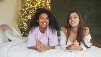 Two young women sit on bed in pajamas and talk with Christmas tree in background
