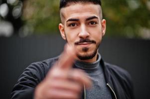 Portrait of stylish arab beard man wear grey turtleneck and black jaket. Arabian model guy show finger close to camera. photo