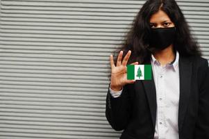 Asian woman at formal wear and black protect face mask hold Norfolk Island flag at hand against gray background. Coronavirus at country concept. photo