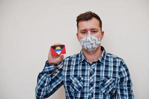 Man in checkered shirt show Antigua and Barbuda flag card in hand, wear protect mask isolated on white background. American countries Coronavirus concept. photo