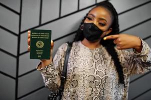 mujer africana con mascarilla negra muestra el pasaporte de guinea-bissau en la mano. coronavirus en el país africano, cierre de fronteras y cuarentena, concepto de brote de virus. foto