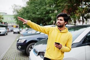 Urban young hipster indian man in a fashionable yellow sweatshirt. Cool south asian guy wear hoodie calling taxi by online app at phone. photo