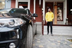 Urban young hipster indian man in a fashionable yellow sweatshirt. Cool south asian guy wear hoodie stand on porch of the house with black business car. photo