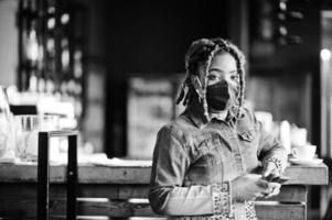 elegante mujer afroamericana con rastas de cabello afro, usa chaqueta de jeans y máscara de protección facial en el restaurante, sostiene el teléfono celular. nueva vida normal después de la epidemia de coronavirus. foto