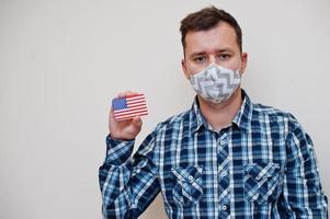 Man in checkered shirt show USA flag card in hand, wear protect mask isolated on white background. American countries Coronavirus concept. photo