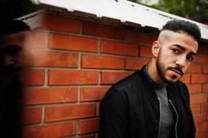 retrato de un elegante hombre de barba árabe con cuello alto gris y jaket negro. chico modelo árabe contra la pared de ladrillo. foto