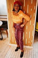 Elegant afro american woman in red french beret, big gold neck chain polka dot blouse and leather pants pose indoor. photo