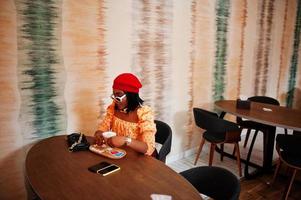 Elegant afro american woman in red french beret, big gold neck chain polka dot blouse pose indoor, sitting at restaurant and drink latte. photo