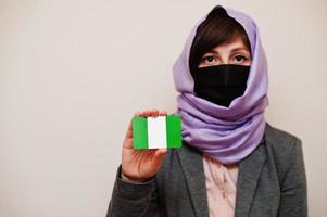 Portrait of young muslim woman wearing formal wear, protect face mask and hijab head scarf, hold Nigeria flag card against isolated background. Coronavirus country concept. photo