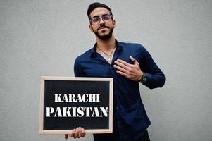 el hombre árabe usa camisa azul y anteojos sostiene una tabla con la inscripción karachi pakistán. ciudades más grandes en el concepto del mundo islámico. foto