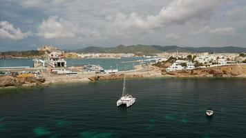 Fly over view of yacht in calm waters near Ibiza with road and buildings in background, a bird flies across the frame video