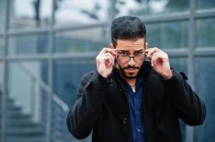 Middle eastern entrepreneur wear black coat and blue shirt, eyeglasses against office building. photo