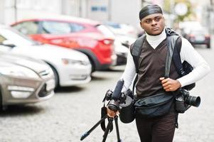 Young professional african american videographer holding professional camera with tripod pro equipment. Afro cameraman wearing black duraq making a videos. photo