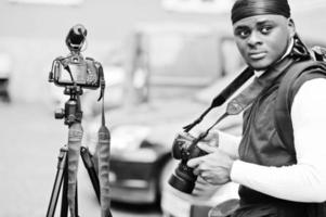 Young professional african american videographer holding professional camera with tripod pro equipment. Afro cameraman wearing black duraq making a videos. photo