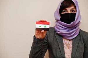 retrato de una joven musulmana que usa ropa formal, protege la máscara facial y el pañuelo de la cabeza del hiyab, sostiene la tarjeta de la bandera siria contra un fondo aislado. concepto de país coronavirus. foto