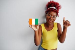 mujer africana con cabello afro, use camiseta amarilla y anteojos, sostenga la bandera de costa de marfil aislada en fondo blanco, muestre el pulgar hacia arriba. foto