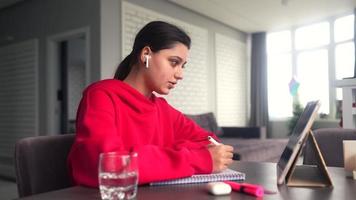 junge Frau in einem hellrosa Sweatshirt sitzt an einem Tisch mit Tablet-Ohrhörern, während sie mit einem Stift in ein Notizbuch schreibt