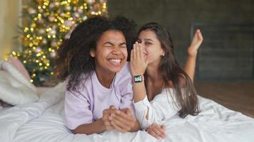 deux jeunes femmes s'assoient sur le lit en pyjama et parlent avec l'arbre de noël en arrière-plan video