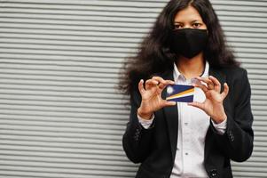 Asian woman at formal wear and black protect face mask hold Marshall Islands flag at hand against gray background. Coronavirus at country concept. photo