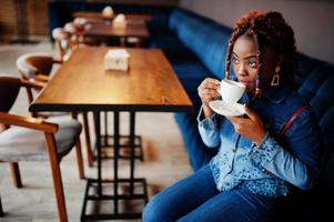 Lovely african american woman with dreadlocks in blue stylish jeans jacket at cafe. Beautiful cool fashionable black young girl indoor drink coffee. photo