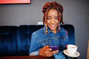 Lovely african american woman with dreadlocks in blue stylish jeans jacket at cafe hold mobile phone. Beautiful cool fashionable black young girl indoor drink coffee. photo