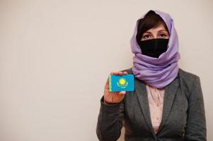 retrato de una joven musulmana que usa ropa formal, protege la máscara facial y el pañuelo de la cabeza del hiyab, sostiene la tarjeta de la bandera de kazajstán contra un fondo aislado. concepto de país coronavirus. foto