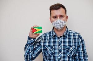 Man in checkered shirt show Guyana flag card in hand, wear protect mask isolated on white background. American countries Coronavirus concept. photo