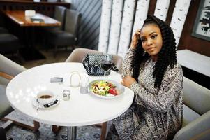 joven mujer afroamericana con estilo sentada en el restaurante, disfrutando de comida saludable con vino.