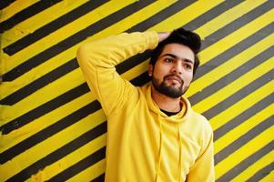 Urban young hipster indian man in a fashionable yellow sweatshirt. Cool south asian guy wear hoodie against striped background. photo