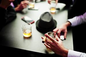 Group of handsome retro well-dressed man gangsters spend time at club, sitting and drinking whiskey. Multiethnic male bachelor mafia party in restaurant. Hand of man with cigar. photo
