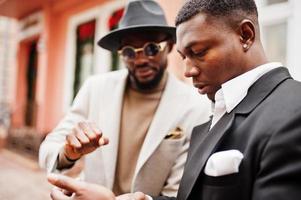 Two fashion black men stand near business car and look at cell phone. Fashionable portrait of african american male models. Wear suit, coat and hat. photo
