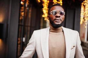 Stylish african american man wear beige jacket with sunglasses pose against house with golden garland. photo