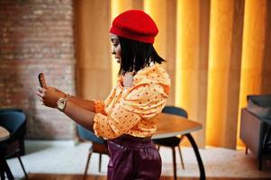 Elegant afro american woman in red french beret, big gold neck chain polka dot blouse and leather pants conducts a live broadcast by mobile phone for social network. photo