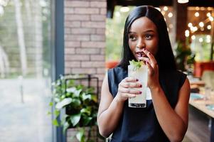 Mujer afroamericana feminista de moda vestida con camiseta negra y pantalones cortos, posada en un restaurante con vaso de limonada. foto