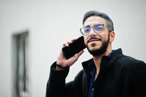 Middle eastern entrepreneur wear black coat and blue shirt, eyeglasses standing against white wall, smoking cigarette and speak by phone. photo