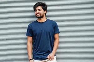 un hombre indio informal con estilo usa una camiseta azul posando contra una pared gris. foto