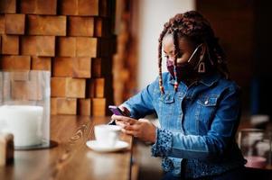 Stylish african american woman with dreadlocks afro hair, wear jeans jacket and face protect  mask at restaurant, hold cellphone. New normal life after coronavirus epidemic. photo