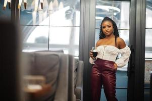 bella mujer afroamericana con blusa blanca y pantalones de cuero rojo posan en el restaurante con una copa de vino. foto