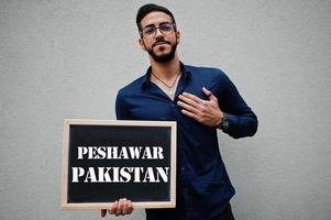 el hombre árabe usa camisa azul y anteojos sostiene el tablero con la inscripción de peshawar pakistán. ciudades más grandes en el concepto del mundo islámico. foto