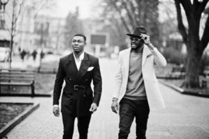 dos hombres negros de moda caminando por la calle. retrato de moda de modelos masculinos afroamericanos. llevar traje, abrigo y sombrero. foto