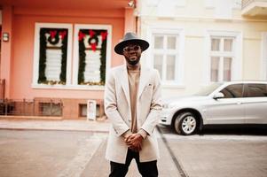 Stylish african american man wear beige jacket and  black hat with sunglasses pose against house with new year decorations and wreath. photo