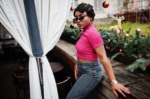 mujer afroamericana joven urbana en top rosa y jeans grises y gafas de sol con bolso. mujeres afro moda chic. foto