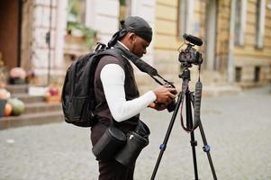 Young professional african american videographer holding professional camera with tripod pro equipment. Afro cameraman wearing black duraq making a videos. photo