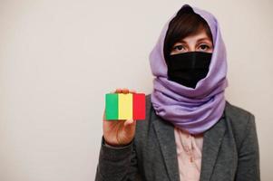 Portrait of young muslim woman wearing formal wear, protect face mask and hijab head scarf, hold Mali flag card against isolated background. Coronavirus country concept. photo