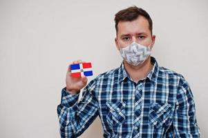 Man in checkered shirt show Dominican Republic flag card in hand, wear protect mask isolated on white background. American countries Coronavirus concept. photo